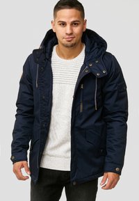 Marineblå parkajakke med hette, frontlynlås, gylne detaljer og flere lommer. Stoffet ser jevnt ut med en slitesterk tekstur.