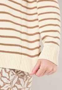 Beige gestrickter Pullover mit horizontalen braunen Streifen. Verfügt über gerippte Ärmelbündchen und Saum. Nahaufnahme des Ärmeldetails, Hand ruht auf dem Saum.