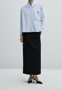 Chemise bleu clair à manches longues avec un détail de poche à volants, associée à une jupe midi noire et des escarpins noirs à bout pointu. Texture du tissu lisse.