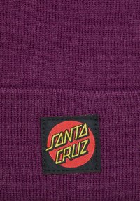 Gorro de punto morado con textura acanalada, que presenta un parche rectangular negro con el logo "SANTA CRUZ" en rojo y amarillo.
