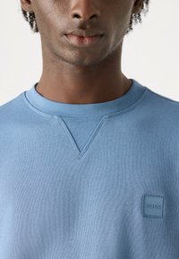 Sudadera de color azul claro hecha de algodón, con cuello redondo y un pequeño parche de logo cuadrado en el pecho. Textura suave, diseño informal.