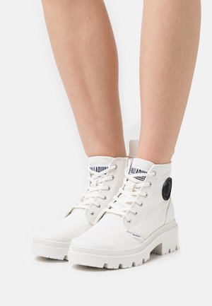 Białe sneakersy za kostkę z płótna i grubymi gumowymi podeszwami. Posiadają czarne akcenty z logo oraz kontrastujące białe sznurowadła.