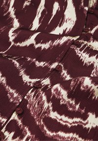 Bordeauxrotes und weißes gemustertes Gewebe mit einem wellenartigen Design, versehen mit braunen Knöpfen und einer strukturierten Oberfläche. Das Design erzeugt einen Schichteffekt.