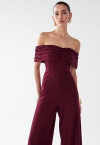 Bordeauxroter Jumpsuit mit schulterfreiem Ausschnitt, gerafftem Stoffdetail, tailliertem Oberteil und weitem Bein. Hergestellt aus glattem, dehnbarem Material.