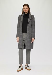 Grijs geblokte jas met inkepingen, twee knopen en lange mouwen, gedragen over een zwart coltrui en grijze broek. Schoeisel heeft crèmekleurige en bruine accenten.