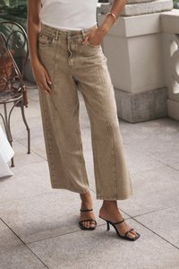 Šviesiai rudos plačios kelnes su priekinėmis siūlėmis, aukštu liemeniu ir dviem priekinėmis kišenėmis, derinkite su juodais aukštakulniais ant lygios, plytelėmis išklotos paviršiaus.