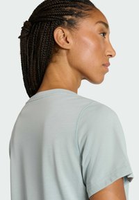 Mladá osoba s vrkočmi, ktorá má na sebe svetlosivú košeľu s krátkym rukávom, zobrazená zozadu a zboku na jednoduchom pozadí.