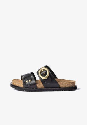 Sandale noire à semelle en liège, deux brides en cuir, dont l'une avec une grande boucle hexagonale dorée, et une semelle extérieure en caoutchouc noir texturée.