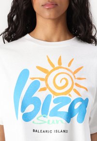 Λευκό βαμβακερό T-shirt με πολύχρωμο σχέδιο που έχει "Ibiza Sun" σε μπλε και πράσινο, γραφικό ήλιου σε πορτοκαλί και κείμενο "Balearic Islands".