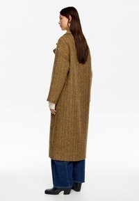 Abrigo largo marrón con patrón de espiga, mangas anchas, tela texturada y un ajuste ligeramente oversize, combinado con jeans de pierna ancha en azul oscuro y botas negras.