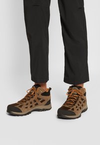 Brązowe buty trekkingowe z siateczkowymi panelami, teksturowanym zamszem i gumowymi akcentami, z pomarańczowymi sznurówkami i średnim krojem dla wsparcia kostki.