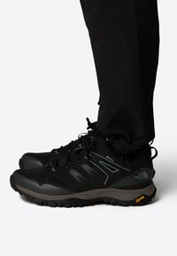 Mustat vaelluskengät tekstuurilla varustetusta verkkopinnasta, vahvistetusta varpaasta ja kumipohjasta, jossa on Vibram-tarkistuksia. Narukiristys ja tukeva muotoilu.