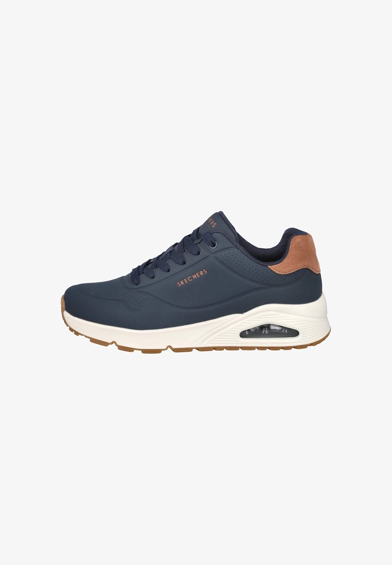 Zapatilla Skechers de color azul marino con talón de gamuza marrón, suela blanca con amortiguación visible y cordones azul marino sobre un fondo blanco.