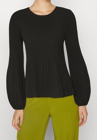 Top lungo a maniche nere a coste con scollo tondo, con fondo svasato e tessuto strutturato, abbinato a pantaloni verdi.