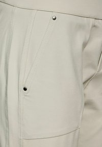 Ljusgrå byxor har en slät yta, snedställda framfickor och tryckknapplås, med synliga sömdetaljer.