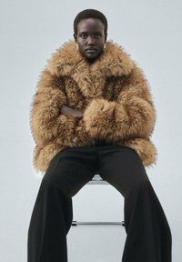 Brown faux fur coat with a wide collar and textured surface, paired with black wide-leg trousers. Model seated on a clear chair.