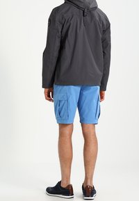 Grå hættetrøje med logo på ærmet, kombineret med blå cargobukser med sidelommer og sorte sneakers med brune detaljer.