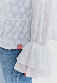 Biała przezroczysta bluzka z teksturowanymi kropkami, mankietami z falbankami oraz małą czerwoną metką, noszona z jasnoniebieskimi dżinsami.