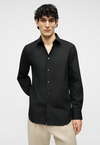 Άνδρας με κοντά σγουρά μαλλιά, φοράει μαύρο πουκάμισο με κουμπιά και μπεζ παντελόνι, στέκεται μπροστά σε μία απλή φωτεινή επιφάνεια.