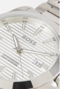 Reloj de acero inoxidable plateado con una esfera blanca rayada, manecillas plateadas y display de fecha. Resistente al agua hasta 50 metros. Logo de la marca centrado.