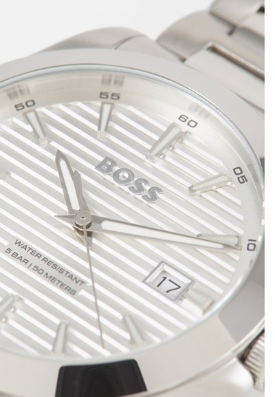Montre en acier inoxydable argenté avec un cadran blanc rayé, aiguilles argentées et affichage de la date. Résistante à l'eau jusqu'à 50 mètres. Logo de la marque centré.