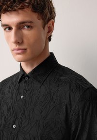 Schwarzes Hemd mit Kragen und Blumenstickerei, mit strukturiertem Stoff, durchgehendem Knopfverschluss und dezentem Glanz. Nahaufnahme der Details des Hemdes.