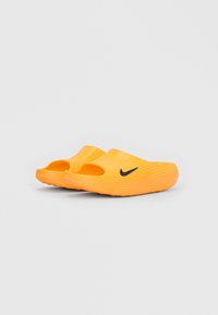 Lyse orange slip-on træsko med tekstureret design og sort Nike swoosh-logo på siden, placeret på ensfarvet lyst baggrund.