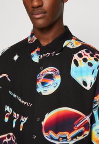 Camicia a maniche corte in tessuto nero con stampe grafiche colorate di dadi, ruote e testo, con colletto a bottoni e dettagli lucidi.