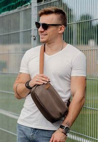 Hnědá crossbody kabelka s hladkým povrchem a jedním popruhem, nošena přes bílé tričko a v kombinaci s džíny. Jsou nasazeny černé sluneční brýle.