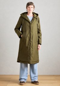 Olijfgroene lange jas met een capuchon, voorzien van drukknoopsluitingen, twee voorzakken en een gewatteerde textuur. Gecombineerd met losse lichtblauwe spijkerbroeken.