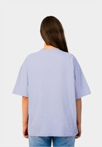 Lichtpaarse oversized t-shirt met korte mouwen en een ronde halslijn, gemaakt van een gestructureerde stof. Gezicht vanuit de achterkant, gecombineerd met blauwe jeans.