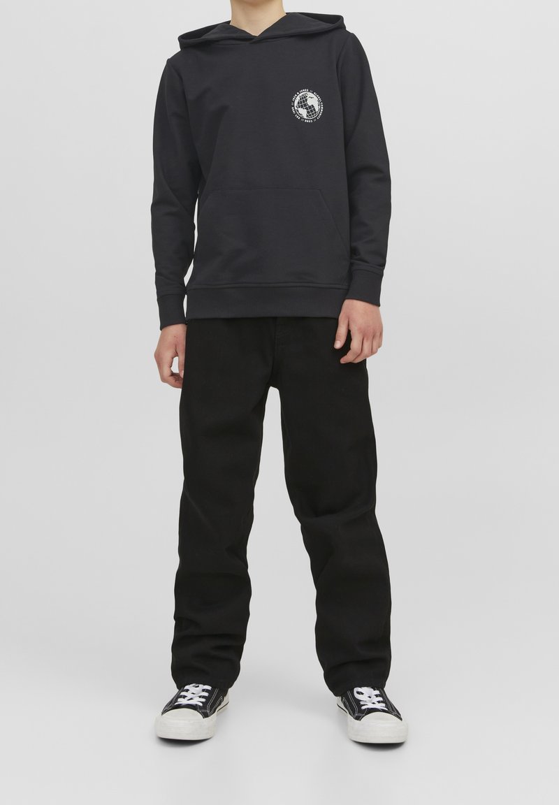 Personne portant un sweat à capuche noir avec un petit logo globe blanc sur la poitrine, un pantalon noir ample et des baskets noir et blanc, debout sur un fond gris.