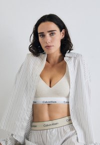 Bež bralette Calvin Klein s V-izrezom, uparen s laganom košuljom preko raznih pruga. Prugasti pojas s detaljem loga.