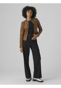 Giubbotto in finta pelle marrone con chiusura a zip, abbinato a pantaloni a gamba larga neri e scarpe argento. Texture liscia, design aderente e dettagli a coste.