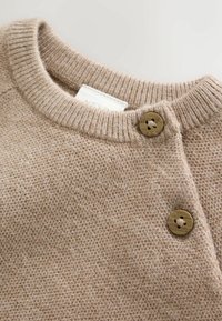 Beige knitted sweater with ribbed round neckline and two brown buttons on the shoulder closure.