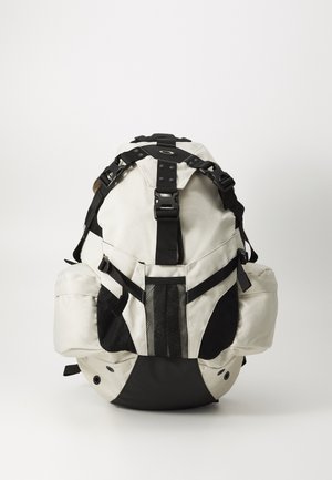 Witte en zwarte rugzak van duurzaam materiaal, met verstelbare schouderbanden, mesh accenten en een ronde basis. Opvallende hardware en ontwerpelementen.