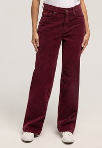 Bordowe spodnie sztruksowe o szerokich nogawkach z fakturowanej tkaniny, z pięcioma kieszeniami i zapięciem na guziki, w połączeniu z białymi trampkami.