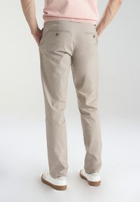 Beżowe spodnie chino o slim fit, z dwiema tylnymi kieszeniami zapinanymi na guziki. W zestawieniu z białymi trampkami i jasnoróżową bluzką.