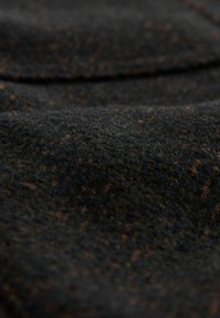 Gros plan d’un tissu foncé texturé avec de fines taches brunes et orange, montrant des fibres douces légèrement surélevées et des plis délicats.