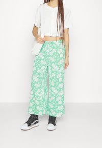 Chemise blanche coupée à boutonnage, pantalon large vert à fleurs, baskets montantes noires et sac à bandoulière blanc. Tissu texturé et coupe décontractée.