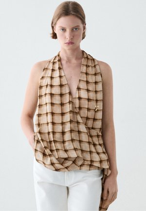 Vrouw met kort haar die een mouwloos beige en bruin geruit haltertopje en een witte broek draagt tegen een effen lichte achtergrond.