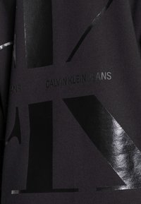 Czarny tekstyl z błyszczącymi czarnymi nadrukami, w tym literami "K" oraz napisem "CALVIN KLEIN JEANS" na ciemnym tle.