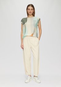 Top à imprimé graphique dans des couleurs pastel, associé à un pantalon crème fuselé et des baskets blanches, avec une coupe décontractée et des manches retroussées.