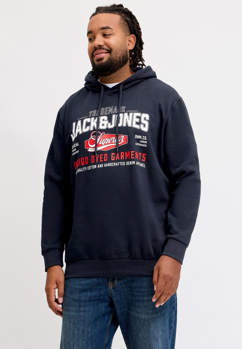Homme avec barbe et dreadlocks portant un sweat à capuche bleu marine Jack & Jones et un jean bleu, debout devant un fond clair uni.