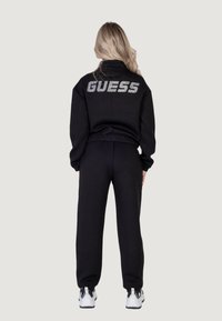 Juodų megztinių ir kelnių rinkinys su trumpu dizainu. Nugarinėje pusėje yra didelis, blizgus "GUESS" logotipas. Tekstūruota medžiaga, laisvas siluetas ir rankogaliai.