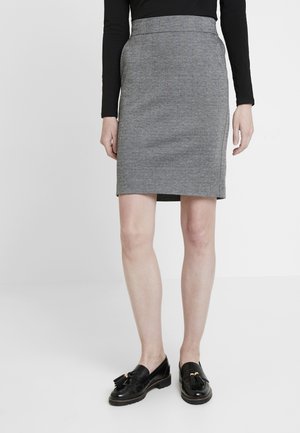 Femme portant une jupe crayon grise mi-genou, un haut noir à manches longues, et des mocassins en cuir noir à pompons, debout sur un sol blanc.