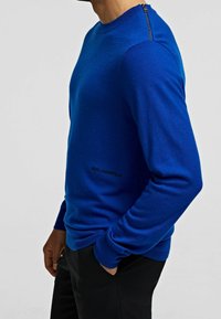 Blaue Strickjacke aus glattem Material mit Rundhalsausschnitt, langen Ärmeln und einem Reißverschlussdetail an der Schulter. Markensymbol an der Seite sichtbar.