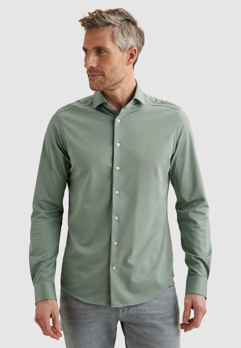 Homme aux cheveux gris portant une chemise ajustée verte sauge à manches longues boutonnée et un jean gris clair, regardant vers la gauche.