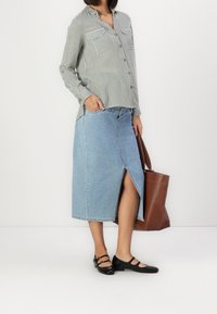 Donna che indossa una camicia a righe con bottoni, una gonna midi in denim azzurro chiaro con spacco frontale, scarpe Mary Jane nere e tiene una borsa a tracolla marrone.