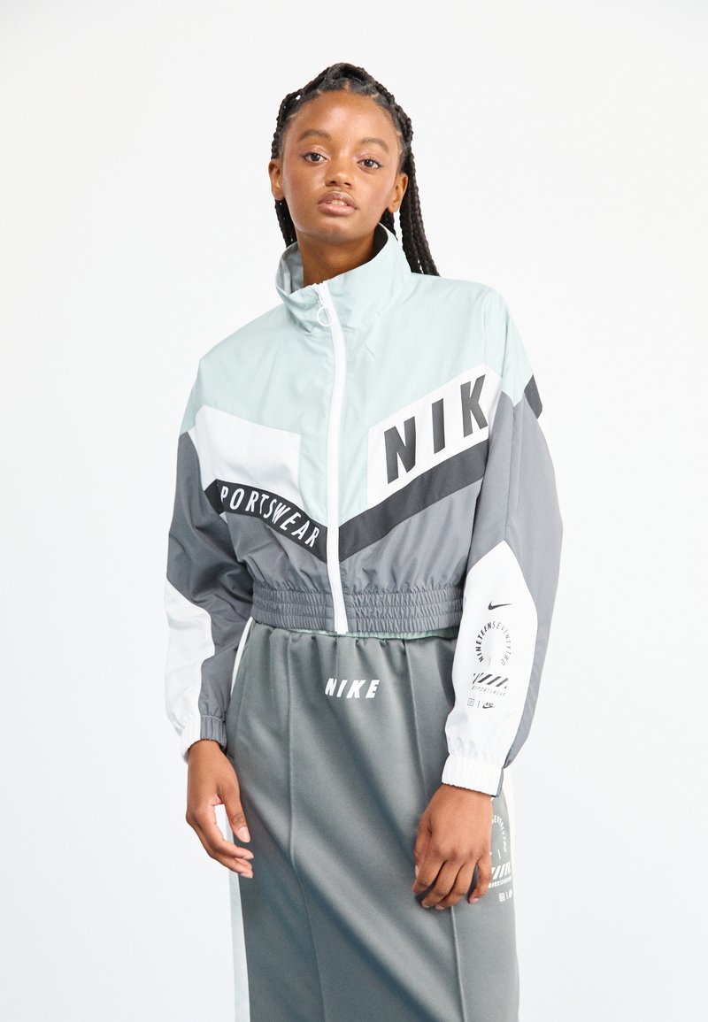 Jachetă sport Nike cu panouri albastre deschise, gri și albe. Previne elastic la talie, închidere cu fermoar și accente cu logo pe mâneci și piept.